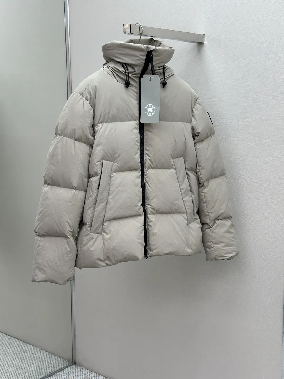 P1010 Canada Goose Black Label Badge Hooded Down Jacket (khaki) The high-quality filling is made of 90 national standard high-quality white duck down, selected high-loft velvet, weighing 240g, light and warm, with excellent temperature locking performance and good resilience. The fabric is woven with 400T high-count yarn. After special water-repellent treatment, the surface forms a lotus leaf effect - raindrops will slide down like beads, and snow particles will fall off when touched. The tear resistance reaches 200N (approximately 20kg load-bearing), the light gray matte texture breaks the stereotype of "outdoor clothing = hardcore", and the soft luster presents a high-end matte effect under urban neon lights, seamlessly switching scenes from ski resorts to CBD office buildings. The ribbon is woven with polyester as the base, and the brand letters are decorated on the ribbon through high-precision computer embroidery. The edges of the pattern are clear, the stitches are fine and even, and multiple layers of embroidery thread are stacked to enhance the three-dimensional effect. Precision thread reinforcement prevents linting and ensures the fluffy and resilient elasticity lasts as new. The embedded cuffs are three-dimensionally woven to form a "wavy" elastic structure, which forms natural folds when fitting the wrist, increasing the contact area with the skin, thus improving the windproof effect. At the same time, micro-velvet treatment is added to the inside of the cuffs, which feels like a second layer of skin and avoids friction on the skin from metal zippers or hard fabrics. Size:S-M-L-XL